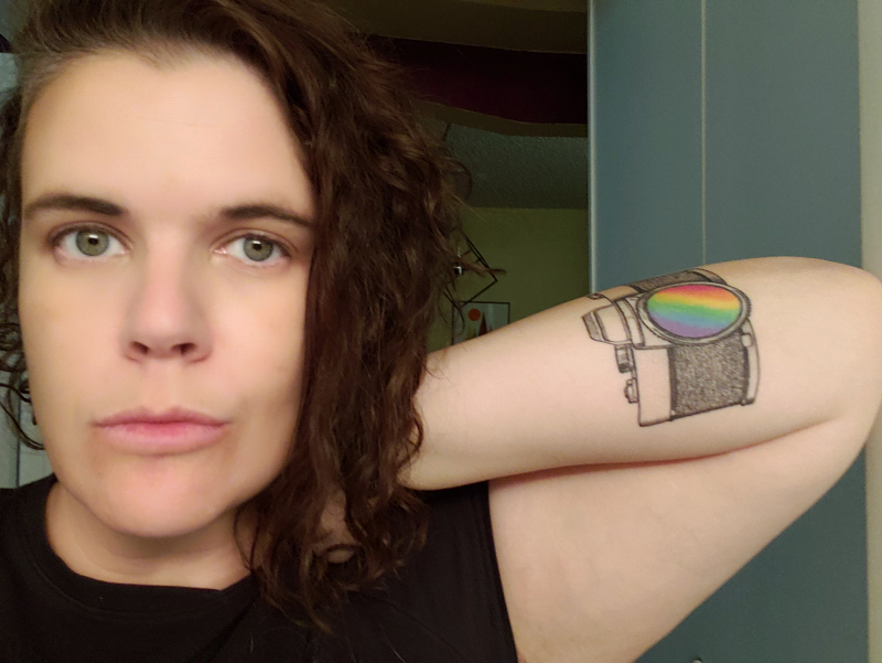 selfie of a white person with curly brown hair side parted on the viewer's left holding their hand behind their head with their elbow leaning against a door frame on the viewer's right. their visible forearm has a tattoo of a 1970s style camera with a rainbow spectrum in the lens