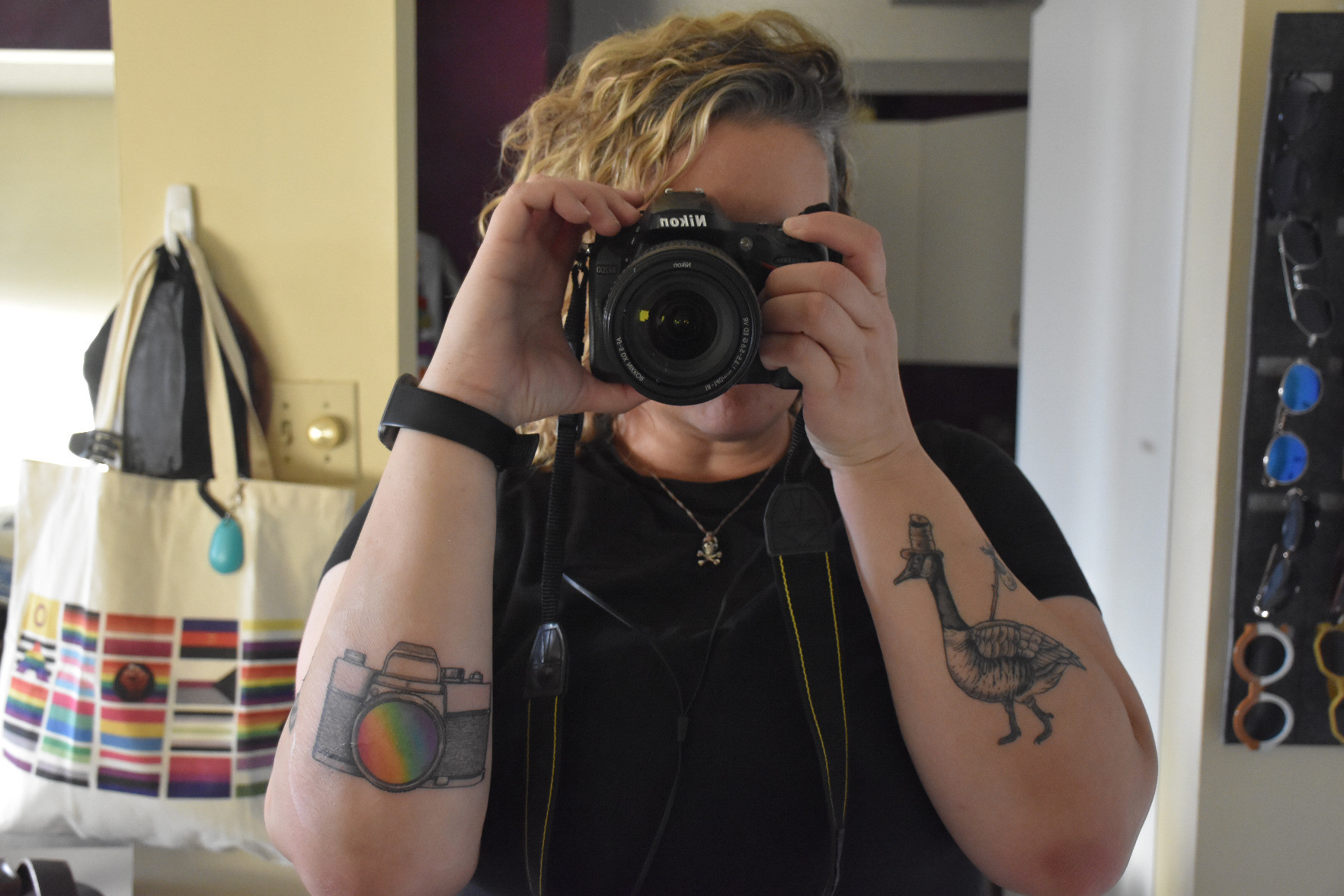 mirror selfie taken with a dSLR that I'm holding up to my face and thus blocking everything but my chin. Wavy/curly blonde hair parted far on the viewer's right is directed towards the viewer's left. On my right arm is a tattoo of the Hobo Goose (a Canada goose with a beat-up hat and bindle on a stick) and on my left arm is a tattoo of a retro SLR with the lens coloured in with a rainbow gradient. The background is mostly clutter because I am a fucking mess.