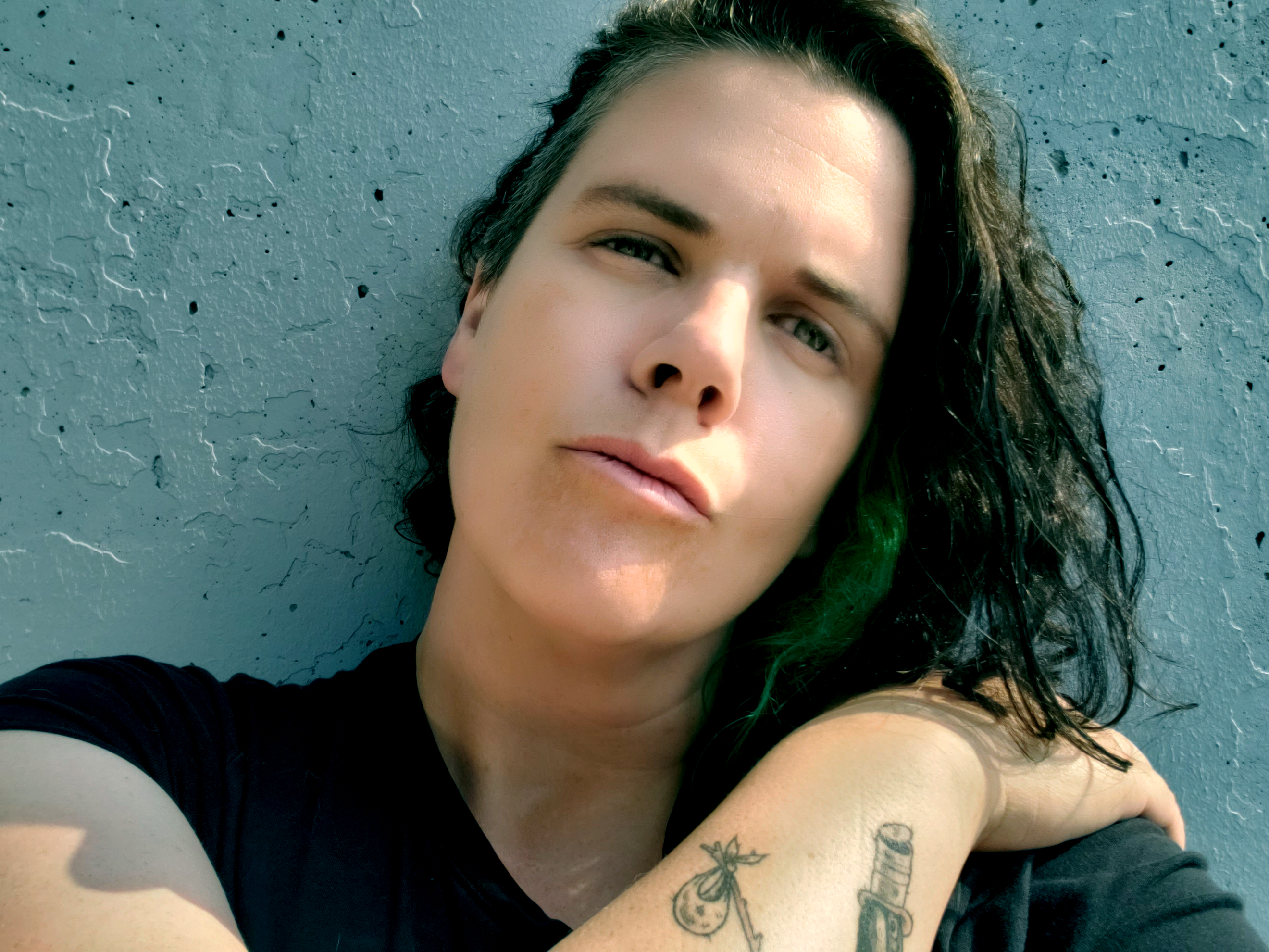 selfie of a white person with curly brown hair side parted on the viewer's left standing against a grey concrete wall holding their left hand over their right shoulder (showing the top of a Hobo Goose tattoo on the wrist) and leaning their head back a bit