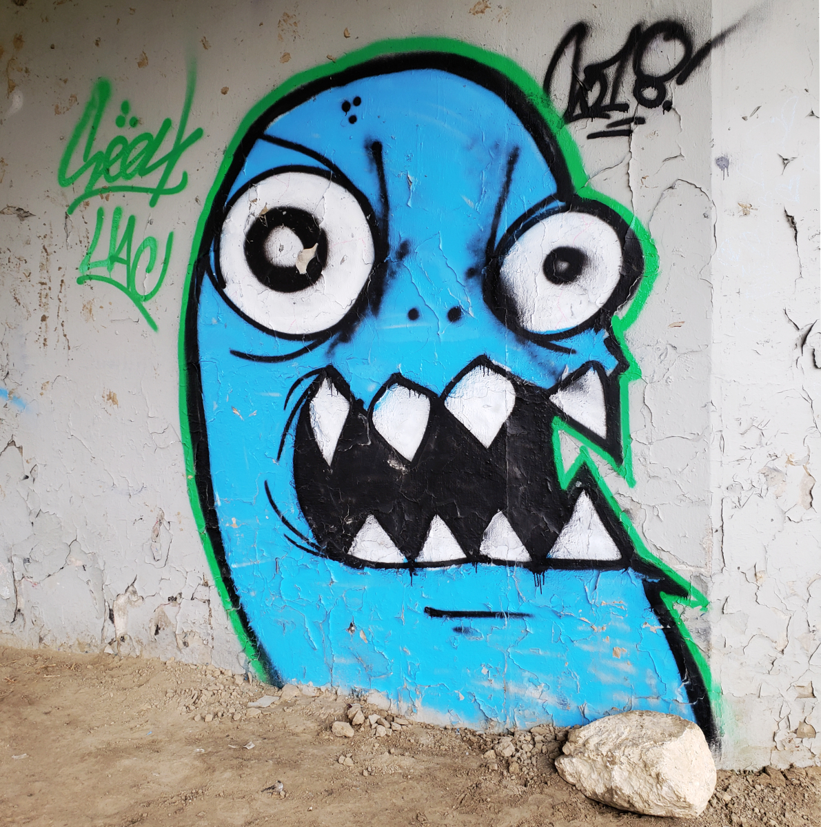 a big blue cartoonish monster with sharp teeth and big eyes with uneven pupils spraypainted on a wall along a dirt path