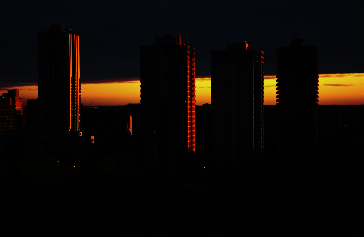 a dark skyline of four high rise apartment buildings against a fiery sky with dark clouds at the top