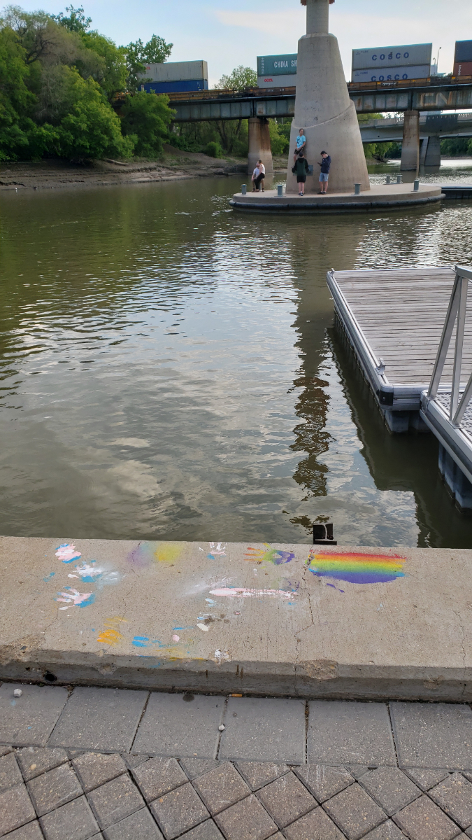 the curb of a pier with handprints and other drawings in trans flag or rainbow pride flag colours. there is a dock on the right side going into a brownish river, with a concrete platform in the midground with young people standing on it. in the background is a railway bridge with a train going by