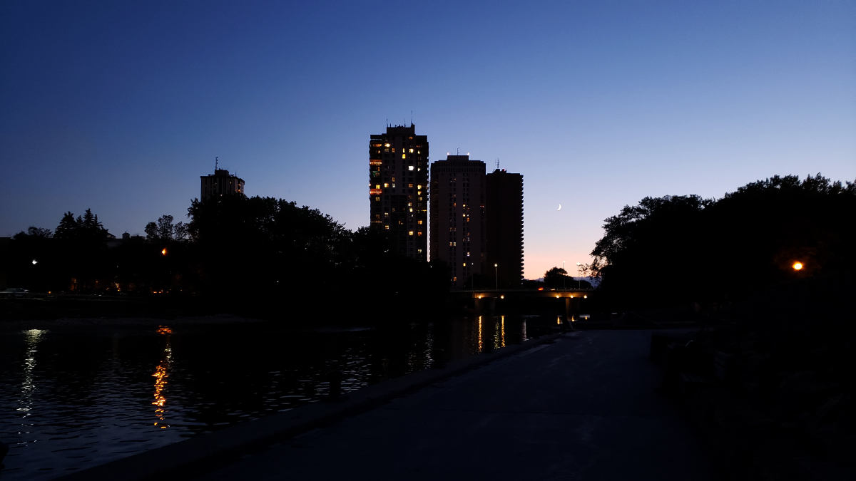 a nighttime shot of the river reflecting lights from high rise buildings in the distance. You can see a crescent moon in a part of the sky that is lighter than the rest