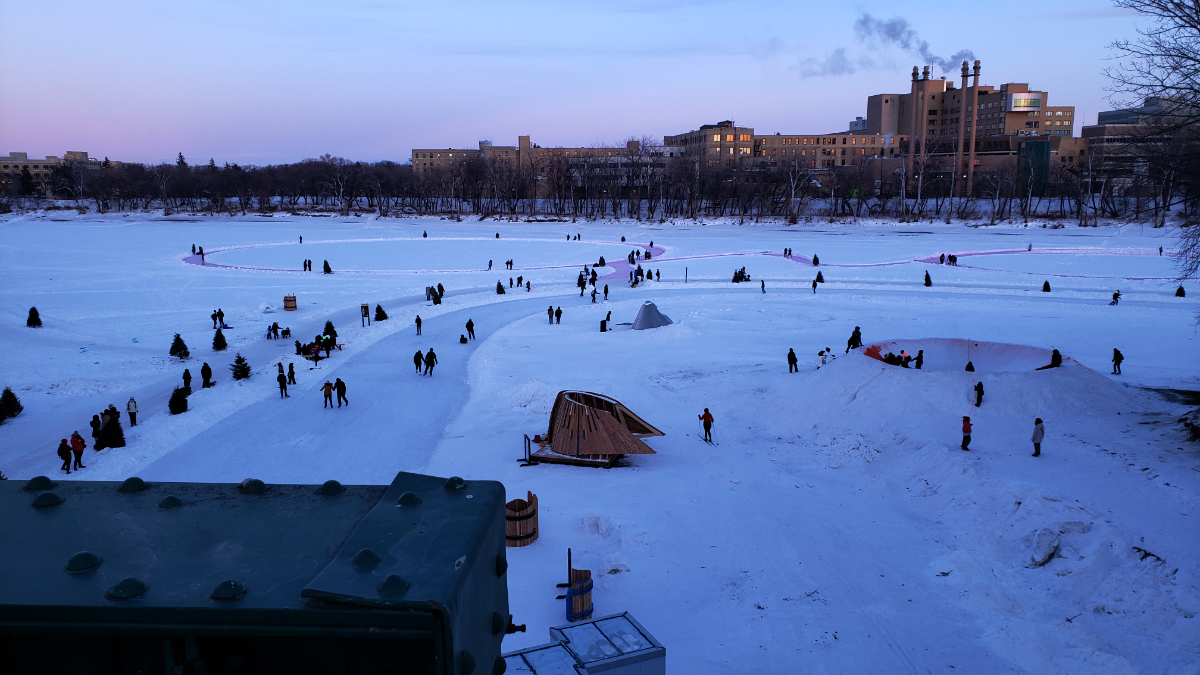 a shot from above of an intersection of two rivers frozen over with snow, where part of the river is changed into an ice path that curves around. dozens of people are walking or skating on the river, which also has different artistic warming huts on it, as well as a crater with orange spray paint to make it look like a volcano