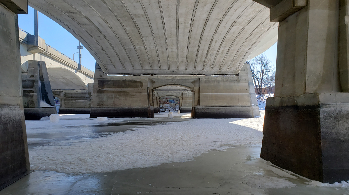 a frozen river from a path beneath a wide concrete bridge. you can see the other side of the bridge through three sets of concrete pillars, where there is colourful graffiti on the wall