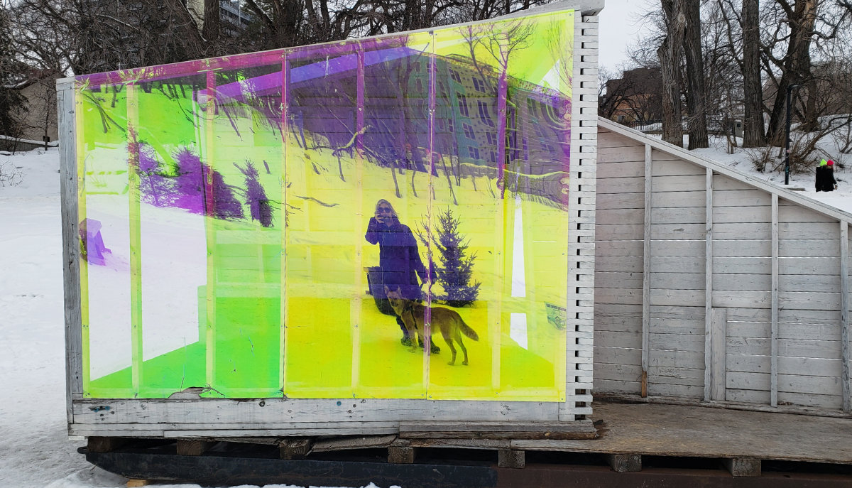 a warming hut on a frozen river with plastic walls of electric citrus colours (green and yellow) that reflect the photographer, in a parka with blonde curly hair and sunglasses, and their dog on a leash looking into the reflection. In the background there are people walking on a higher path on the riverbank.