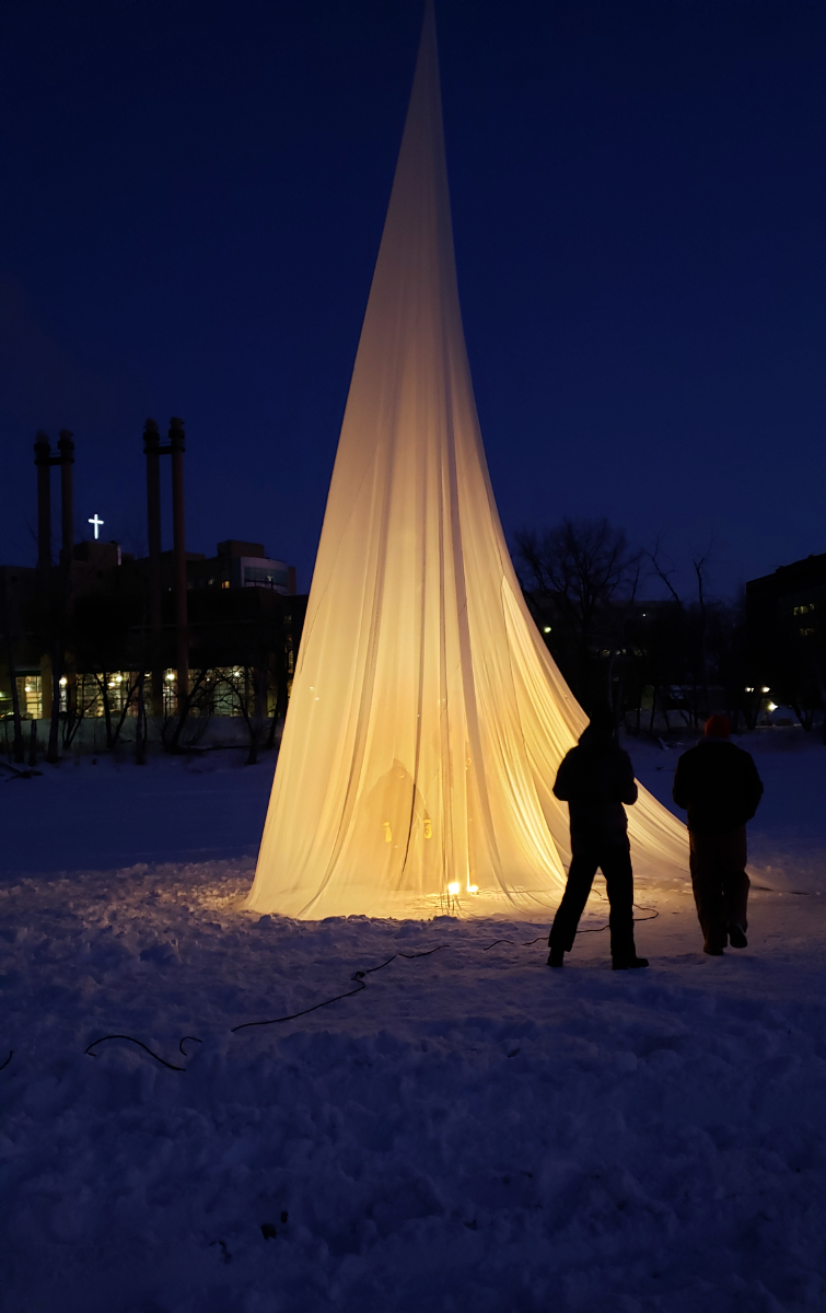 a nighttime shot of a tent made of a light cloth draped over a pole with a light shining from within it, placed on top of snow on a frozen river. you can see the silhouette of two people standing between the photographer and the tent. in the background you can see barren trees and chimneys from buildings