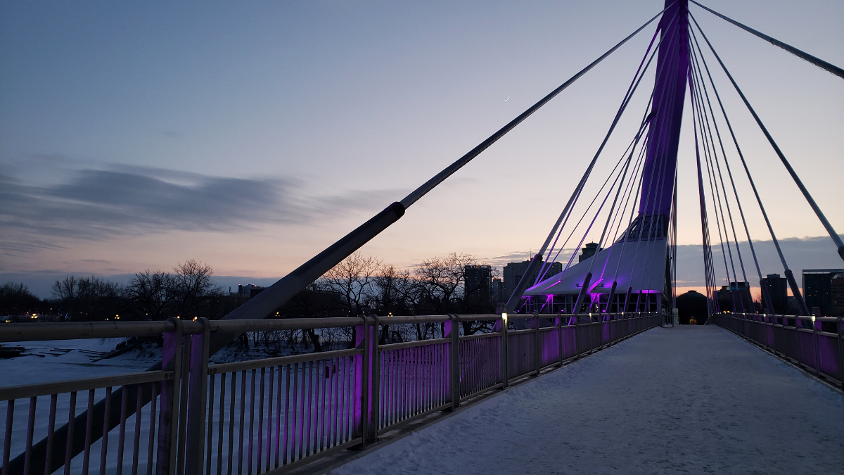 a pedestrian bridge covered in snow over a frozen river at dusk. The river has cables going up to a tall pole with a small kiosk-like structure on it. The lights coming from below the bridge are a bright purple. There are barren trees and modest cityscapes in the background