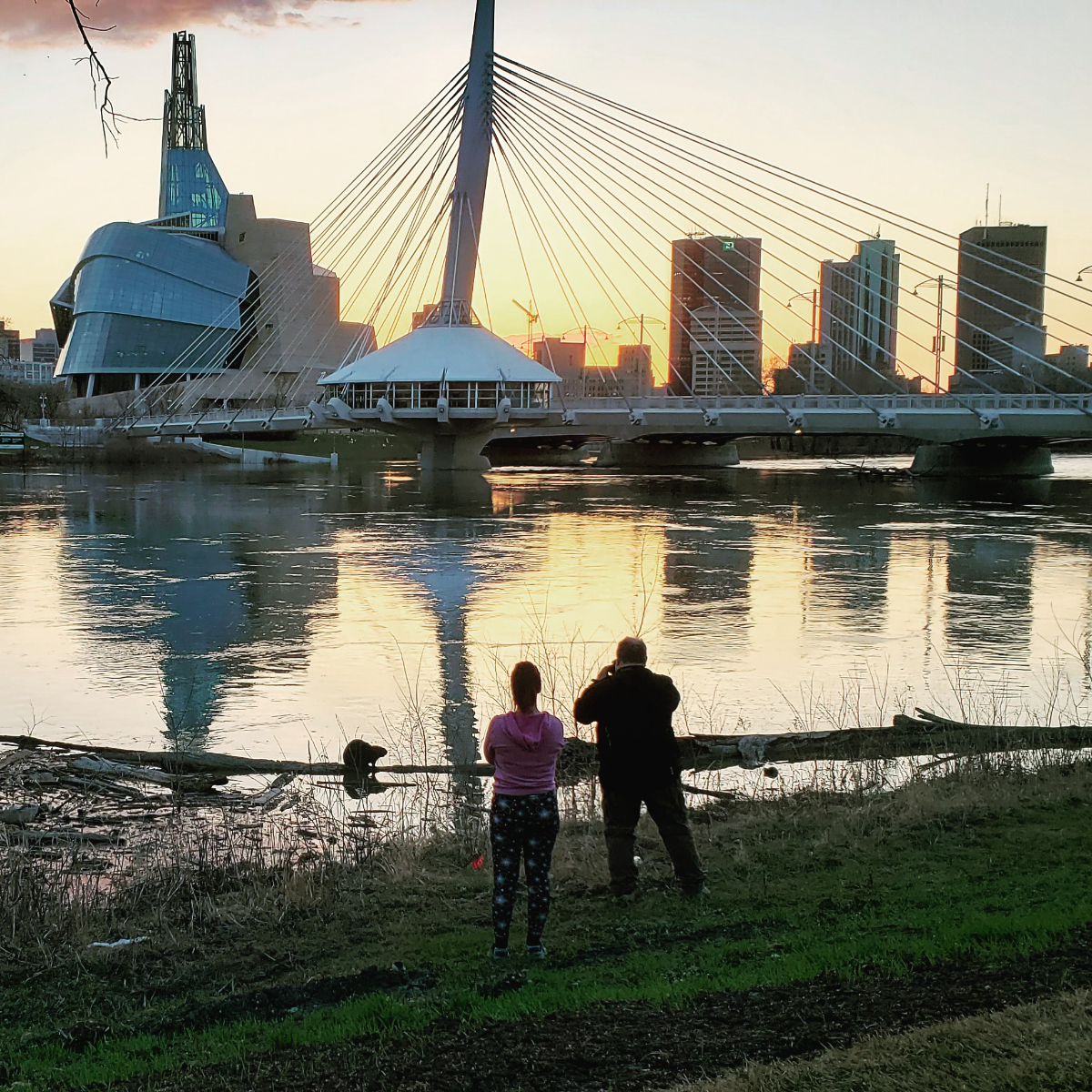 a background of the sun setting behind a Winnipeg skyline featuring the Canadian Museum of Human Rights (a funky looking huge building), the Esplanade Riel (a bridge with a tall spike and suspension cables coming from it), and the tall towers of Portage and Main. The foreground is the Red River, where two people are standing on the banks taking a photo of a beaver who is sitting on a log.