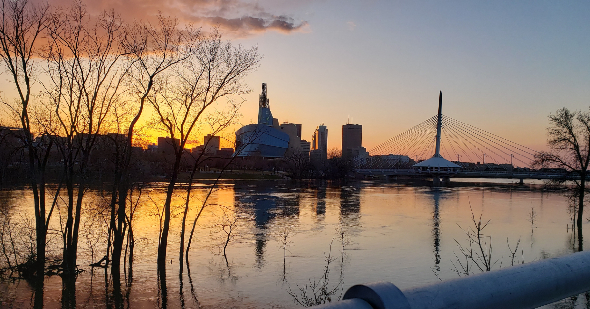 a river at sunset with a dark orange horizon and the silhouette of a flowing shaped building on the other side with a bridge to the right