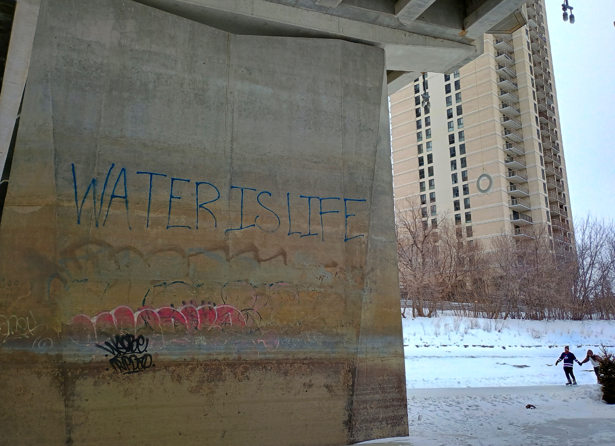 A wide concrete support wall under a concrete bridge on a snow-covered icy river. The wall has a variety of graffiti, but most notably 'WATER IS LIFE' in large blue letters. In the background is a high rise apartment building, a snowy river bank, a couple ice skating on the river, and a lifesaver hanging down from the bridge.
