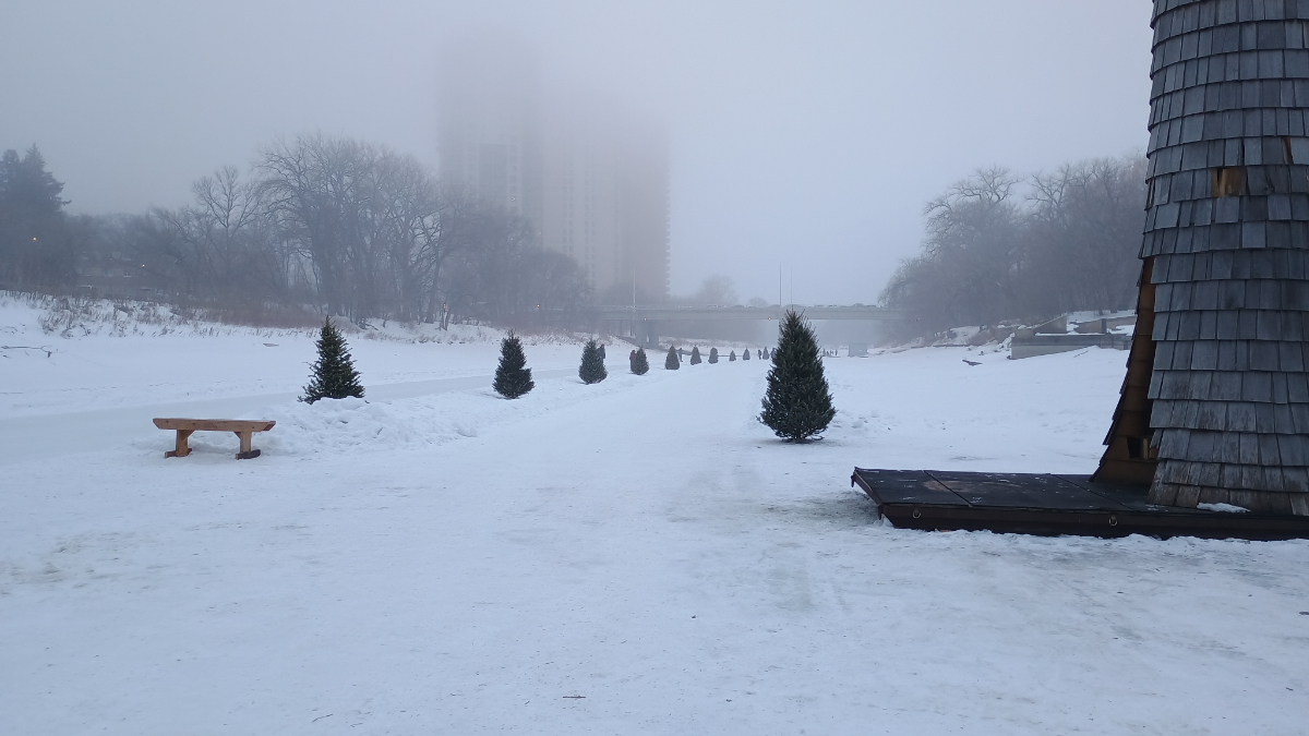 a frozen river surface with a tall shingled warming hut on the right and a fog over a path lined with trees that heads towards/under a bridge in the distance
