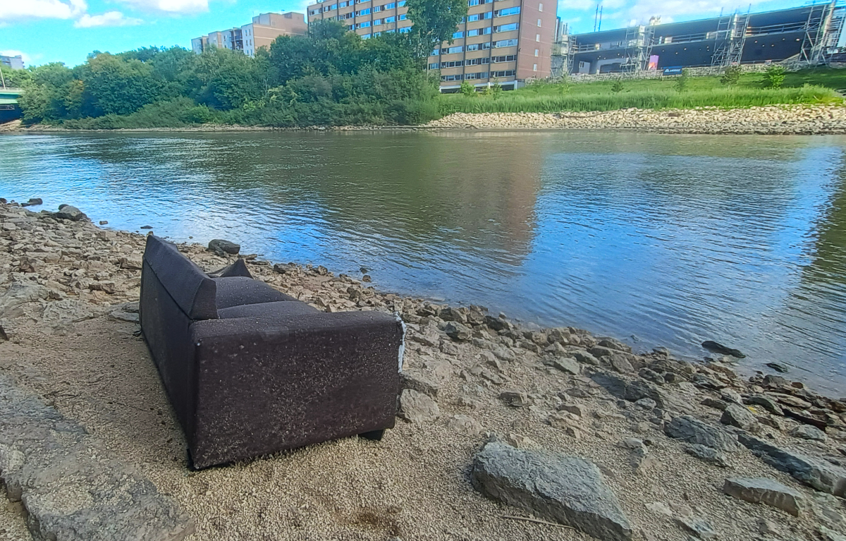 a brown loveseat sitting on sands and rocks alongside the Assiniboine River. on the opposite side there is a wide apartment block and another one in construction that is just for now concrete levels