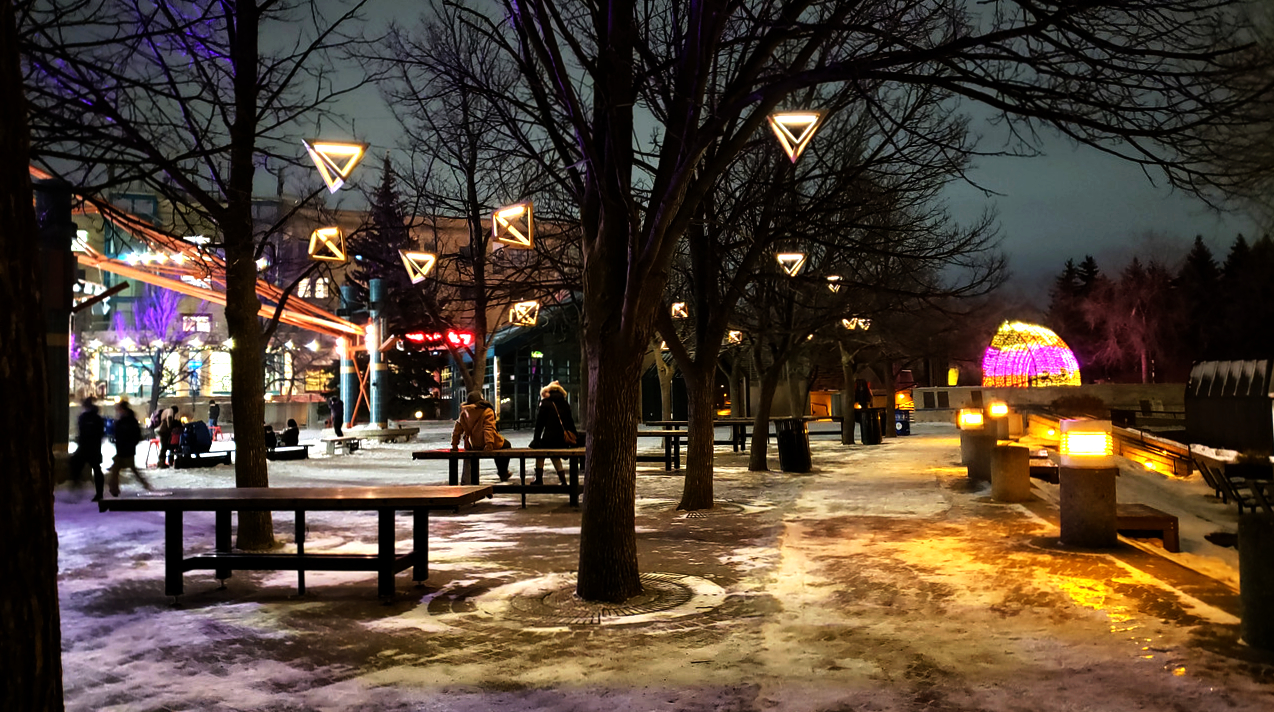 a winter nighttime scene of a public area with tables, triangular lights hanging from trees, people walking around, people sitting and putting on/taking off skates, and an arched passageway with pink and yellow lights in the distance. There isn't any water here but I assure you it's right by the frozen river trail.