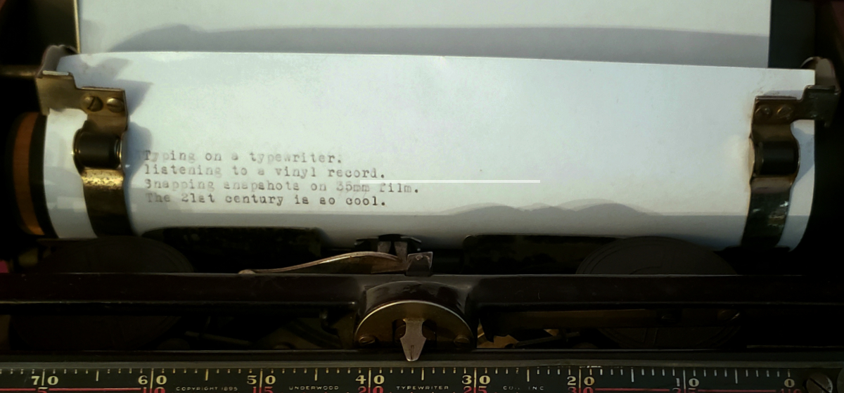 a piece of paper in an old school typewriter saying: Typing on a typewriter. / listening to a vinyl record. / Snapping snapshots on 35mm film. / The 21st century is so cool.