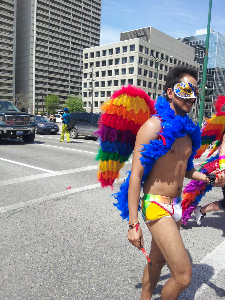 a man in tight rainbow underwear wearing rainbow feathered wings, a blue feather boa, and one of those fancy gala masks covered in blue and gold sequens walking on a street and smiling at someone off camera