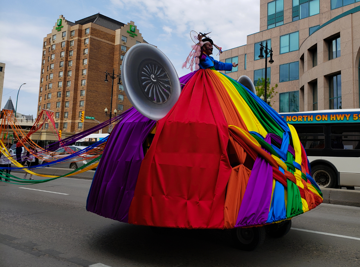 a male performer in fancy theatrical makeup holding a dreamcatcher, wearing a a giant skirt that goes over a cart of some kind moving this person on wheels. the skirt is in rainbow colours and braided at some points. There are strings of fabric from the skirt being held on poles being held by people a couple metres behind this person