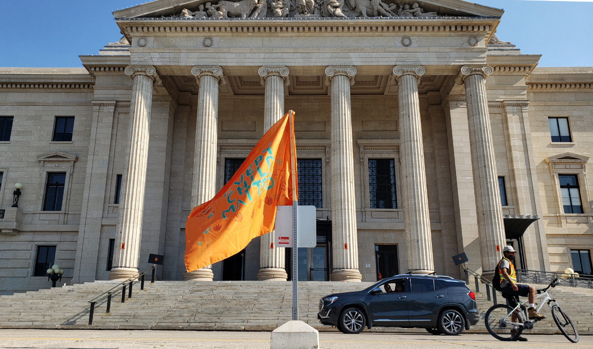 an orange flag with 'EVERY CHILD MATTERS' in written on it in blue flying in front of the Manitoba Legislative building front entrance, with wide sandstone columns holding a roof over an entrance