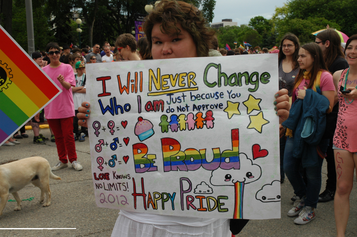 a young person who appears to be Indigenous Turtle Islander holding up a colourful sign saying 'I will never change who I am just because you do not approve. Be Proud. Happy Pride. Love knows no limits 2012.' with colourful drawings of stick figures, gender sign combinations, and a cupcake.