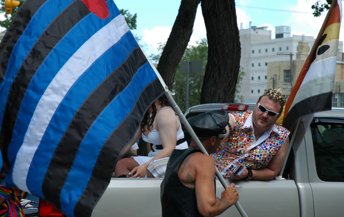 a tan man in a leather vest wearing a leather cap holding a large BDSM pride flag standing next to a truck talking to a burly man sitting in the truck bed and leaning to the side a wearing a colourful bejeweled vest, sunglasses, and a crown, with a bear pride flag next to him