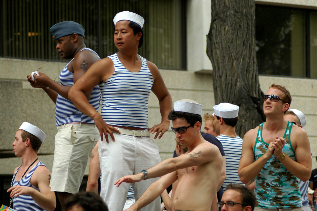 a bunch of men standing on a float dressed as sailors, with nautically striped tank tops, sailors' hats, and fake anchor tattoos on their arms