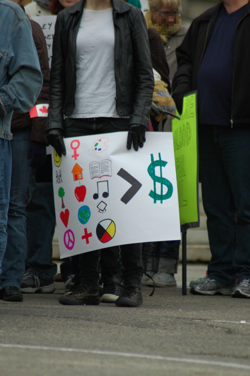 part of a crowd of protesters cropped from the shoulders down, one of whom is holding a sign with a number of symbols: smiley face, venus, paint palette, stick figure family, house, book, tree, apple, musical notes, heart, earth, graduation cap, peace sign, plus sign, medicine wheel; these are next to a greater than sign with $ next to it