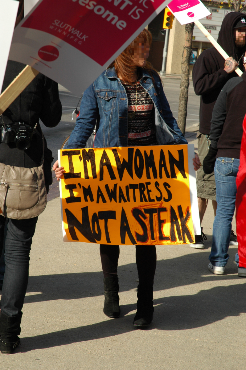 a couple people around the edges carrying professionally printed 'Slut Walk Winnipeg' signs, with a woman standing in the middle with her own handwritten sign saying 'I'm a woman, I'm a waitress, not a steak'