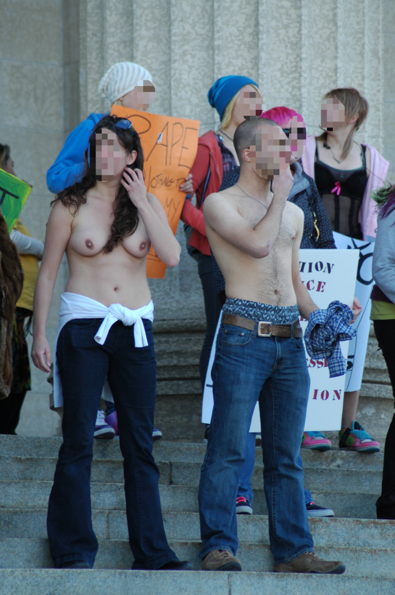 a shirtless woman with long brown hair and a blurred out face on the left, with a shritless bald man with a blurred out face to her right. behind them are blurred face female protesters carrying signs, but the only visible word is 'RAPE' at the top of one of them.