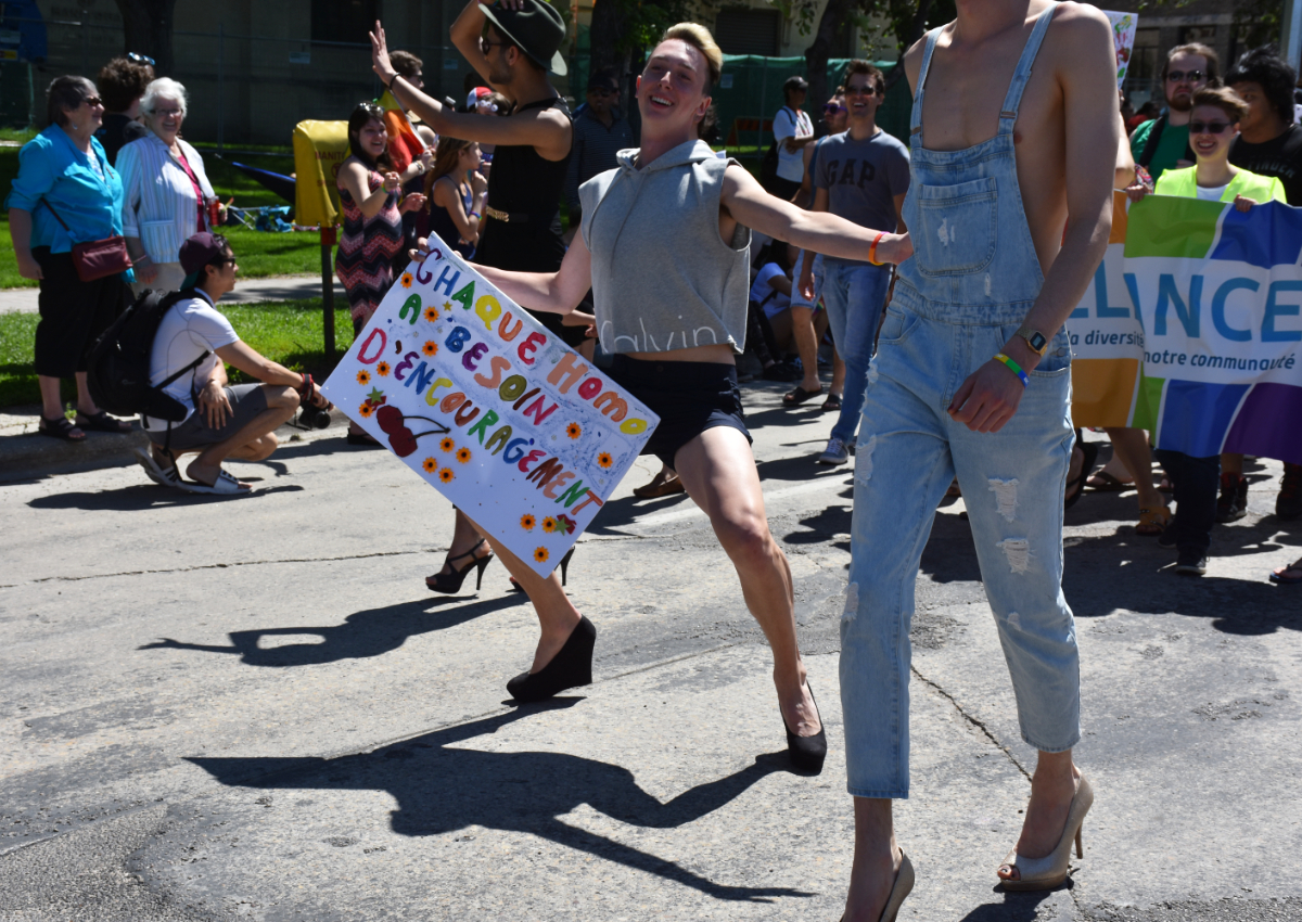 a background of parade onlookers watching a float led by three white men in high heels. one with stubble, sunglasses, a black vest, and holding a black hat on his head is waving at the onlookers. another in short black shorts and a high cut calvin klein grey hoodie is posing with open arms holding a sign in one hand saying 'chaque homo a besoin d'encouragement' (each homo needs encouragement, which I could translate ALL BY MYSELF) with flowers and cherries drawn on it. the third guy is very tall so his head is cropped off, but he's wearing light denim overalls and no shirt. there is a group behind them carrying a large banner with some partial words in French.