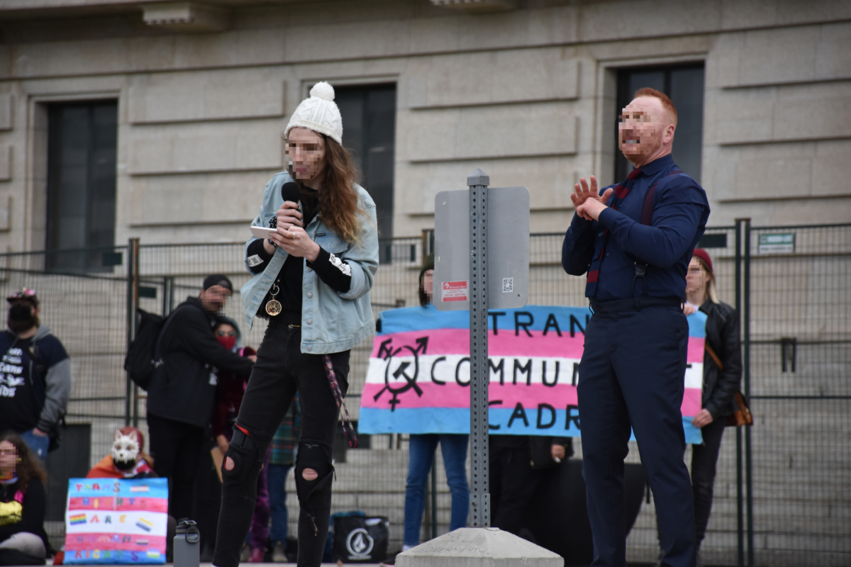 a trans woman with long hair wearing a white toque and a light blue jean jacket with scuffed up black jeans looking at notes from cards in her hand. To the viewer's right is a sign language interpreter. Behind them is a long trans flag banner with a symbol that combines the mars/venus/both trans symbol with the hammer and sickle communist symbol, and text on the banner saying 'tran commun cadr' with letters cut off by the interpreter. There are some other people with smaller signs behind the speaker to the left