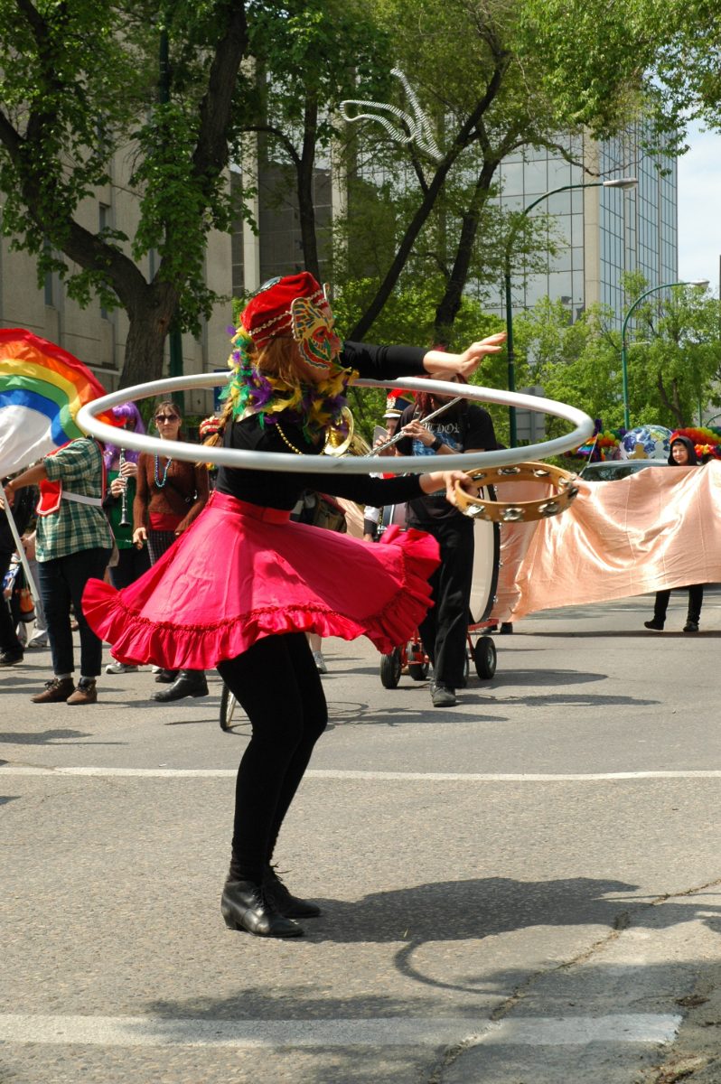 A feminine person with medium length blonde hair wearing a costume mask and hat wearing a black top and tight with a hot pink skirt playing the tambouring while spinning a hoola hoop around their neck