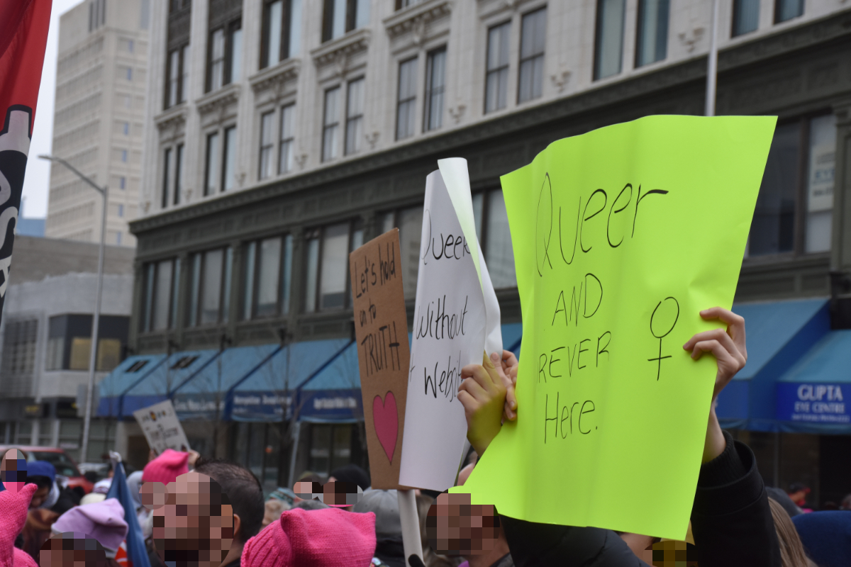 a crowd of people along a busy street with three visible signs: 'Lets Hold Onto Truth' with a heart; 'Queer without website(?)' and 'Queer and FOREVER here' with a venus symbol.