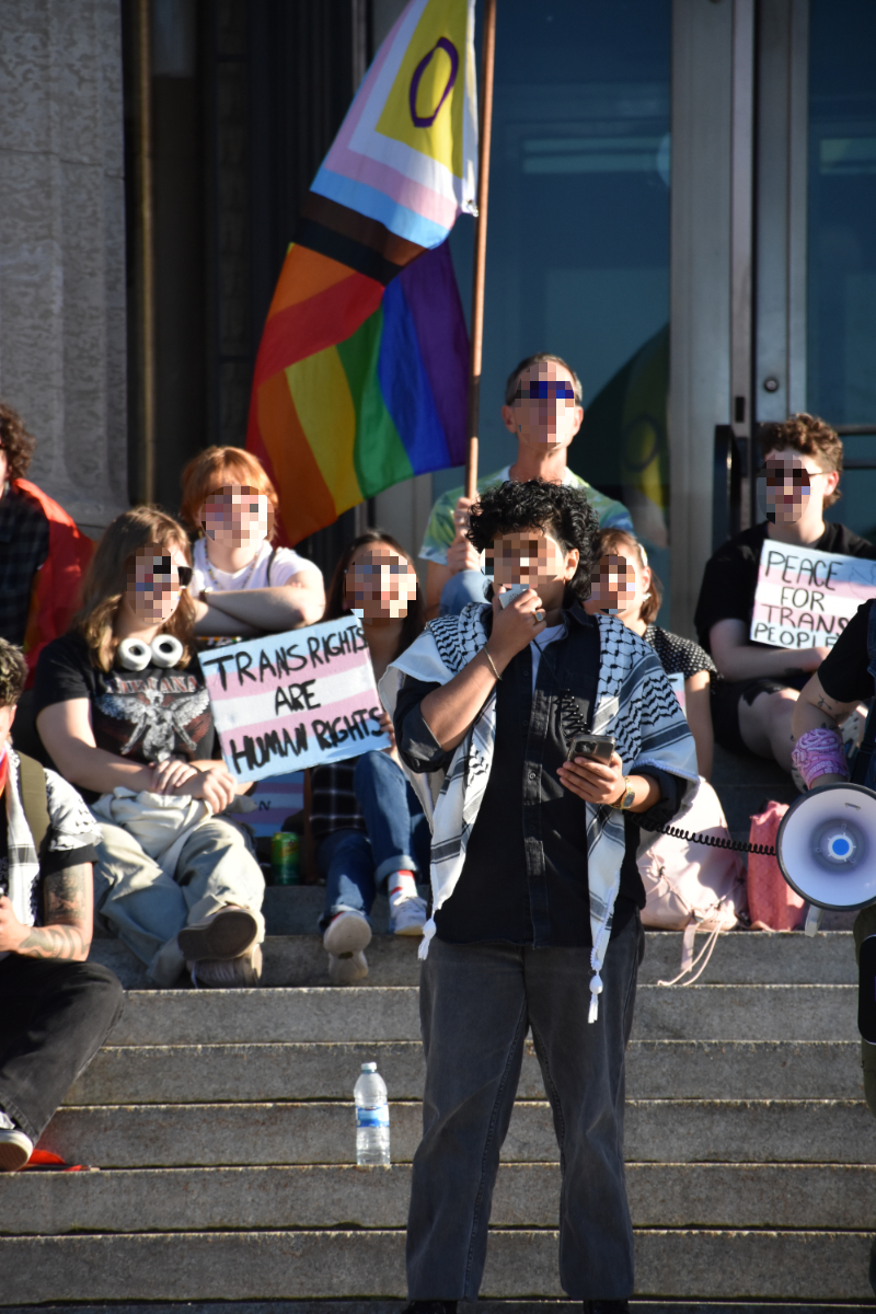 a masculine-presenting person with brown skin and short black curly hair with a black and white keffiyeh over the shoulders on top of a black button up shirt and dark grey jeans holding onto the intercom of a megaphone held by someone to the right. Behind this person are stairs with six other people fully in the shot, carrying signs with the trans flag saying 'Trans Rights are Human Rights' and 'Peace For Trans People' with one person at the very back holding a pole with a large progress pride (intersex version) flag