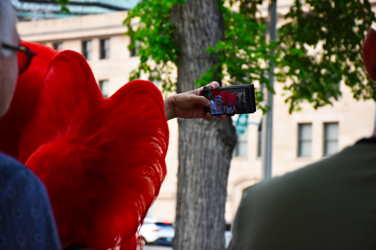 The back of the head of a drag queen wearing a bright red bouffant wig holding a phone taking a selfie in which you can see another drag queen wearing a slightly different bright red bouffant wig posing with her.