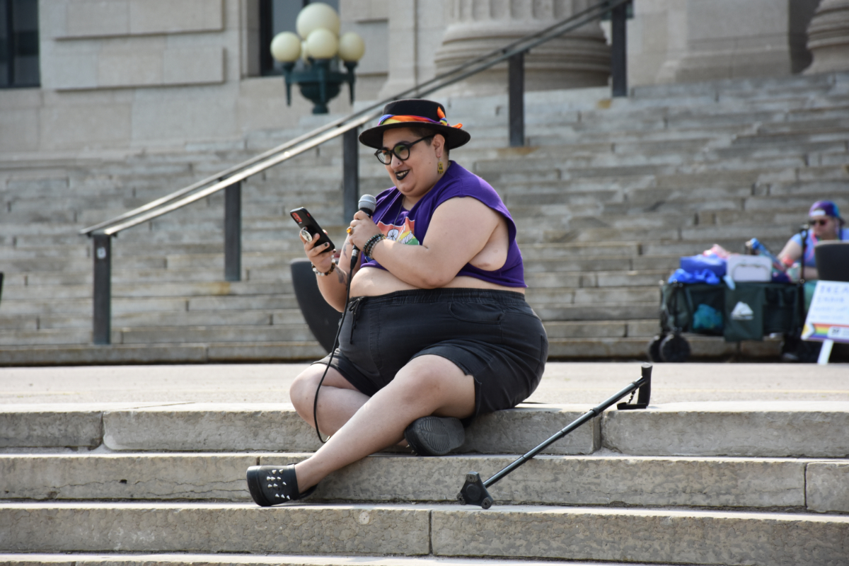 A fat Indigenous queer person sitting on the steps to a legislative building with their cane next to them, looking at their phone and smiling holding a microphone up to their mouth, wearing a very happy outfit with a black hat with colourful decorations, a purple crop top, and black shorts