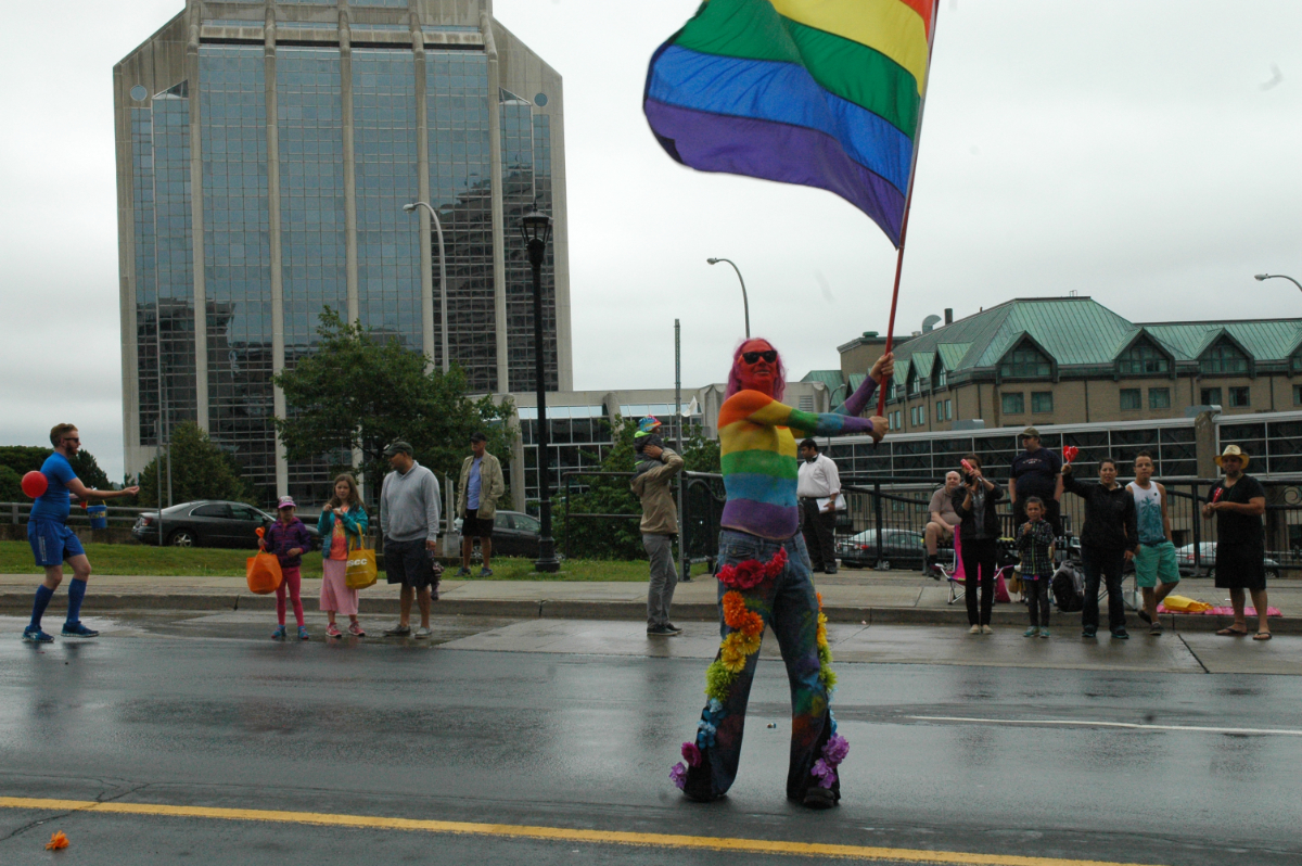 a man painted in rainbow colours from head to waist, wearing bell bottom jeans with rainbow flowers sewn to them. He is holding a big rainbow flag up in the grey sky with a few buildings and a spread out crowd of people in the background.