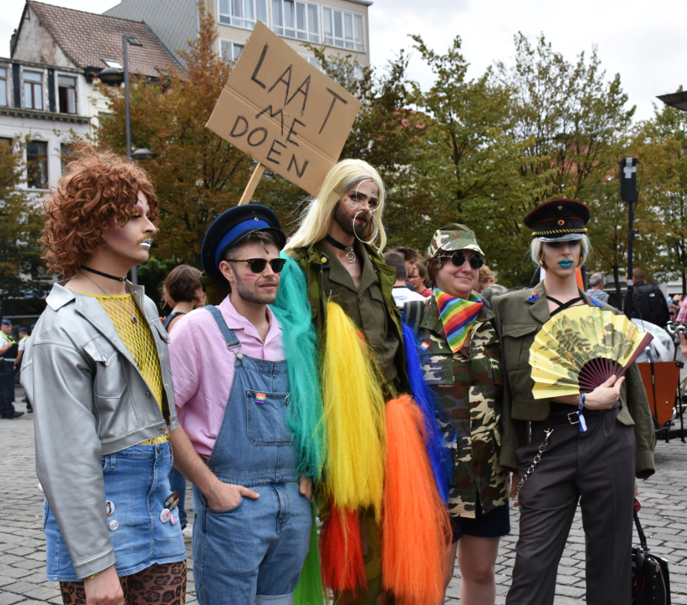 a group of five friends: one masculine person in feminine drag with brown curly hair, silver lipstick, fake eyelashes, black choker, silver vinyl jacket, yellow mesh top, denim skirt with pins on it, and leopart print leggings; one masculine person with a police hat, sunglasses, a light pink golf shirt, and denim overalls rolled up at the knees with pins on the chest; one androgynous tall person with brown facial hair wearing a long straight haired blonde wig, army green clothing, with long tails of synthetic hair in rainbow colours coming from various pockets of the outfit, and large rings coming through a septum piercing with one going up to the hairline and the other down to the chin; one moderately fat butch white person wearing a camouflage hat, sunglasses, a camouflage one-piece, and a rainbow flag; and one thin feminine person wearing an army hat, blonde straight hair (possibly a wig), blue lipstick, a khaki army jacket, and black pants, holding what looks like a Japanese art covered fan over their chest. they are all posing for a photo together with the intended photographer off camera. someone behind them is holding a sign saying 'LAAT ME DOEN' which means 'let me do it' in Dutch/Flemish