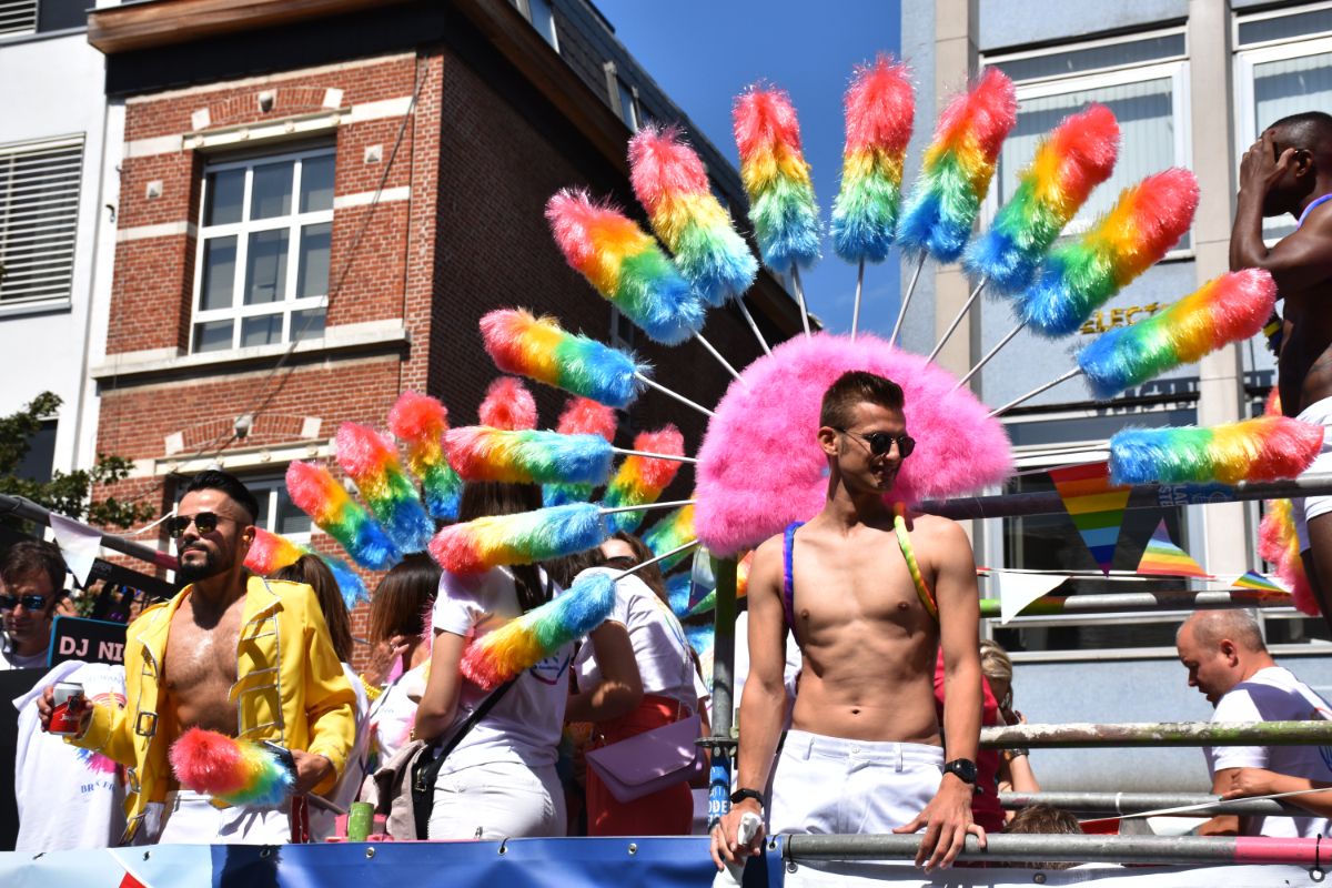 a group standing on the bed of a truck for a pride float, including a white shirtless man of a lean physique wearing something on his back that looks like rainbow feather dusters made into a peacock tail