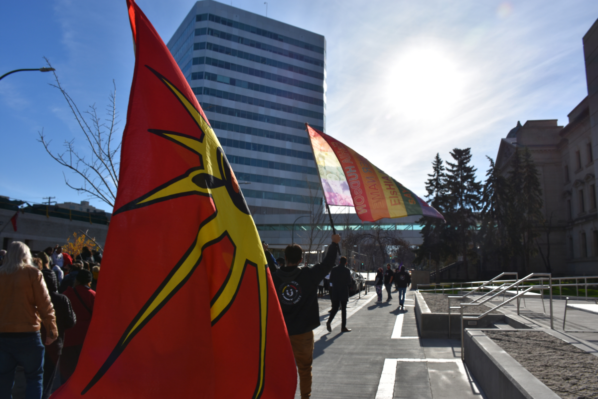 a view from the back right of a parade marching down a street. one person is holding an Indigenous Rights flag (red with a yellow sun-like star with an drawing of an Indigenous person's face, though all you can see here is the adorned feather) and another is holding up a rainbow pride flag with words on it including the name of Eisha Hudson, a 16 year old killed by a police officer.