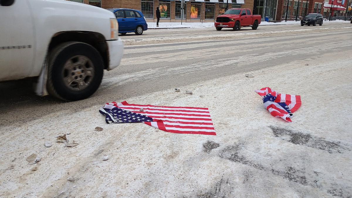 A street with cars, including unnecessarily large pickup trucks probably driven by people who never transport more than their groceries, driving over two US flags that people threw onto the road.