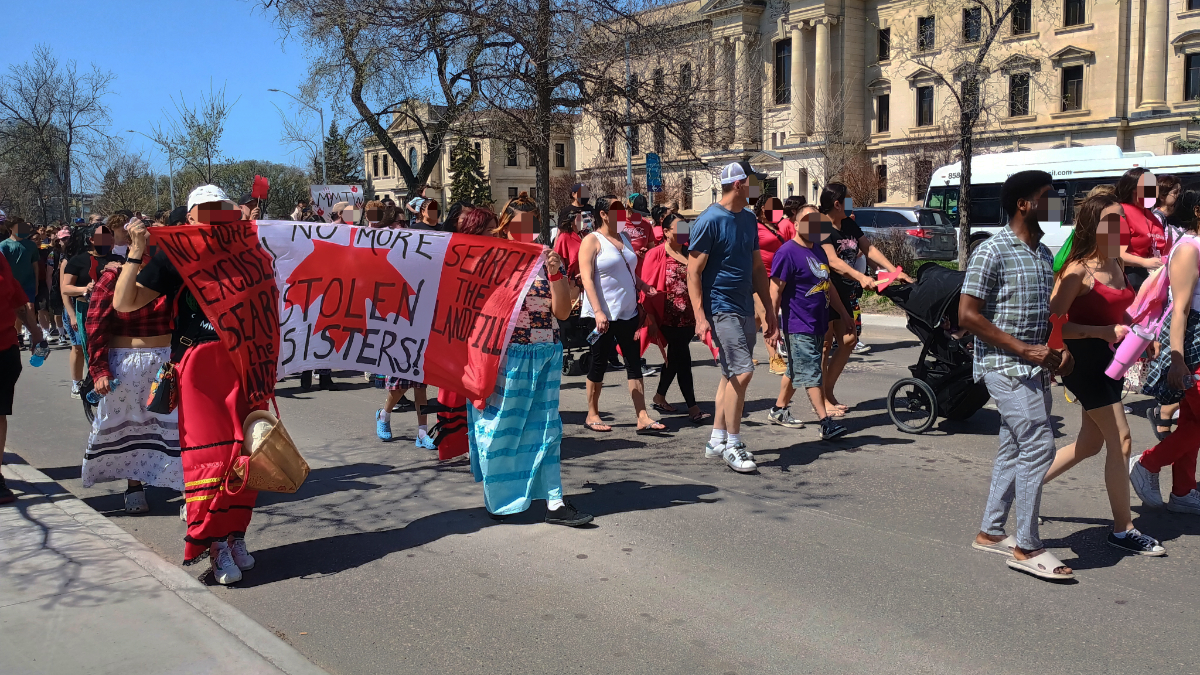 A parade of people, mostly wearing red, of various ages and genders, with blurred out faces, with two women in ribbon skirts holding an upside down Canadian flag saying 'no more stolen sisters'