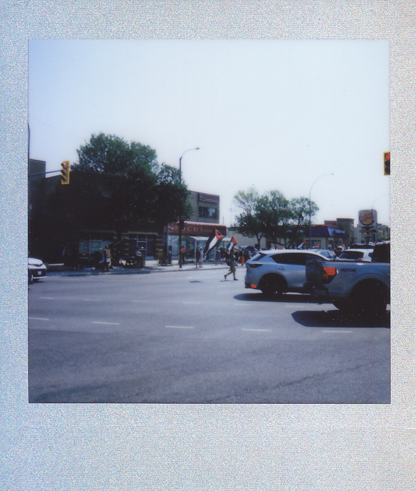 An Instax/polaroid photo of an intersection in the distance with a person carrying a large Palestinian flag while crossing the street, with other people holding Palestinian flags behind them.