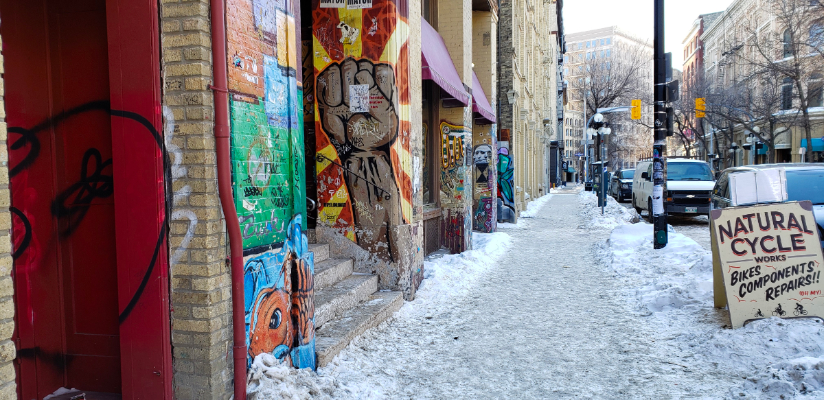 a shot down a street with lots of murals painted on walls outside of building entrances, the most prominent of which is a raised clenched fist with dark skin. The sidewalk is covered in compacted snow and there is slightly more snow with some footprints between the sidewalk and road, lined with parked cars and a sign for a bike store