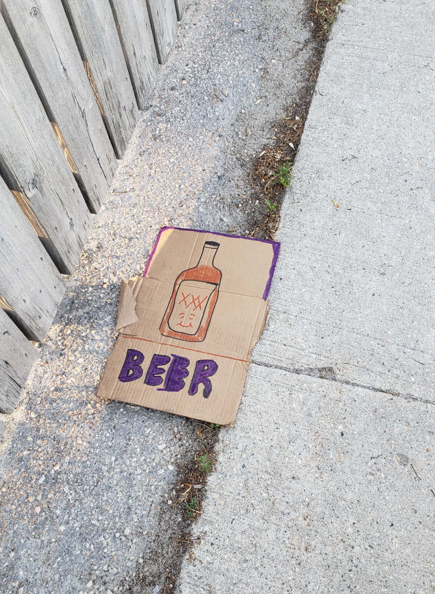 a cardboard sign on the pavement that says 'BEER' in purple letters below a drawing of a smiling bottle that looks more like moonshine than beer, next to a wooden fence