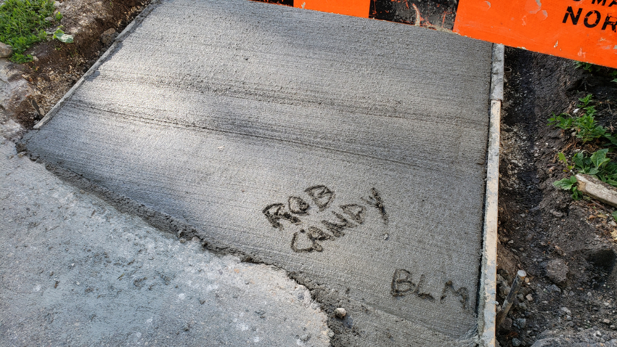 wet cement on a sidewalk with 'ROB CANDY' and 'BLM' freshly written into it