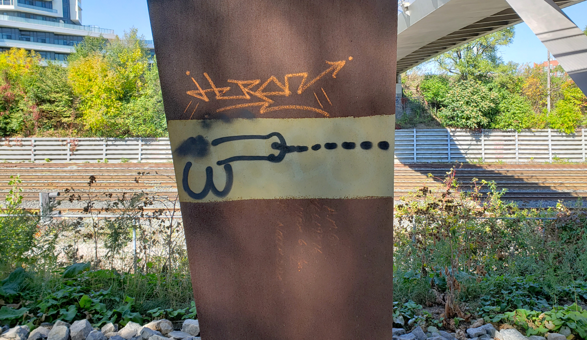 a rusty pillar in front of train tracks and under a bridge to the top left with a load-shooting penis drawn on it in black spray paint