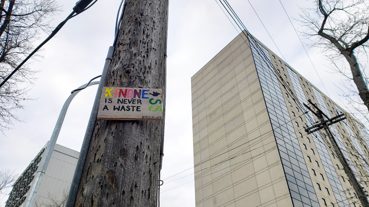 a wooden telephone pole taken from a low angle, with a wood sign nailed to it with the message 'Kindness is Never a Waste' with kindness being written in rainbow colours, and with a black silhouette of a bird after the e, then the ss turn the corner and are at a 90 degree angle from the rest of the message. the sky is cloudy and there are telephone wires and two high rise buildings in the background.