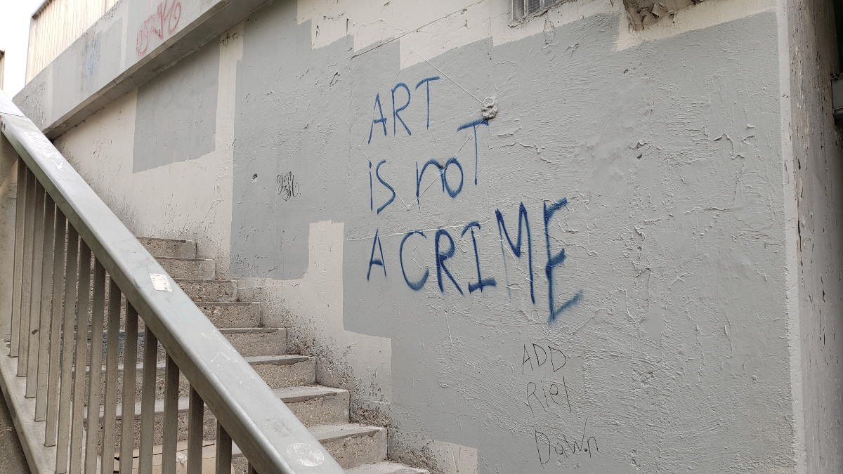 a concrete stairway going up to an overpass with a concrete wall that was painted an off-white colour but now has grey rectangles on it from painting over other graffiti. Part of the grey paint portion has 'ART is not A CRIME' spray painted in blue