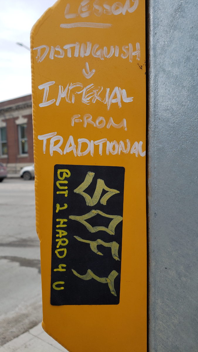 a yellow box attached to a silver streetlight post. There is white handwritten text on the yellow box saying: 'Lesson: Distinguish Imperial from Traditional' and a sideways sticker below it saying 'SOFT but 2 hard 4 u'