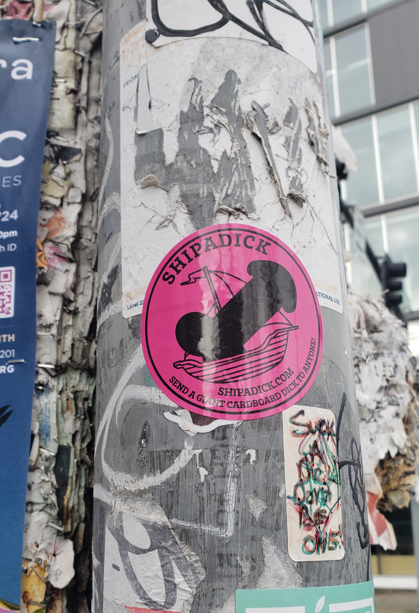 Portland, OR a pole with several stickers, the most prominent of which is round and hot pink with a black image of the shape of a dick on a sailboat with the text arced around it saying 'SHIPADICK send a giant carboard dick to anyone!'